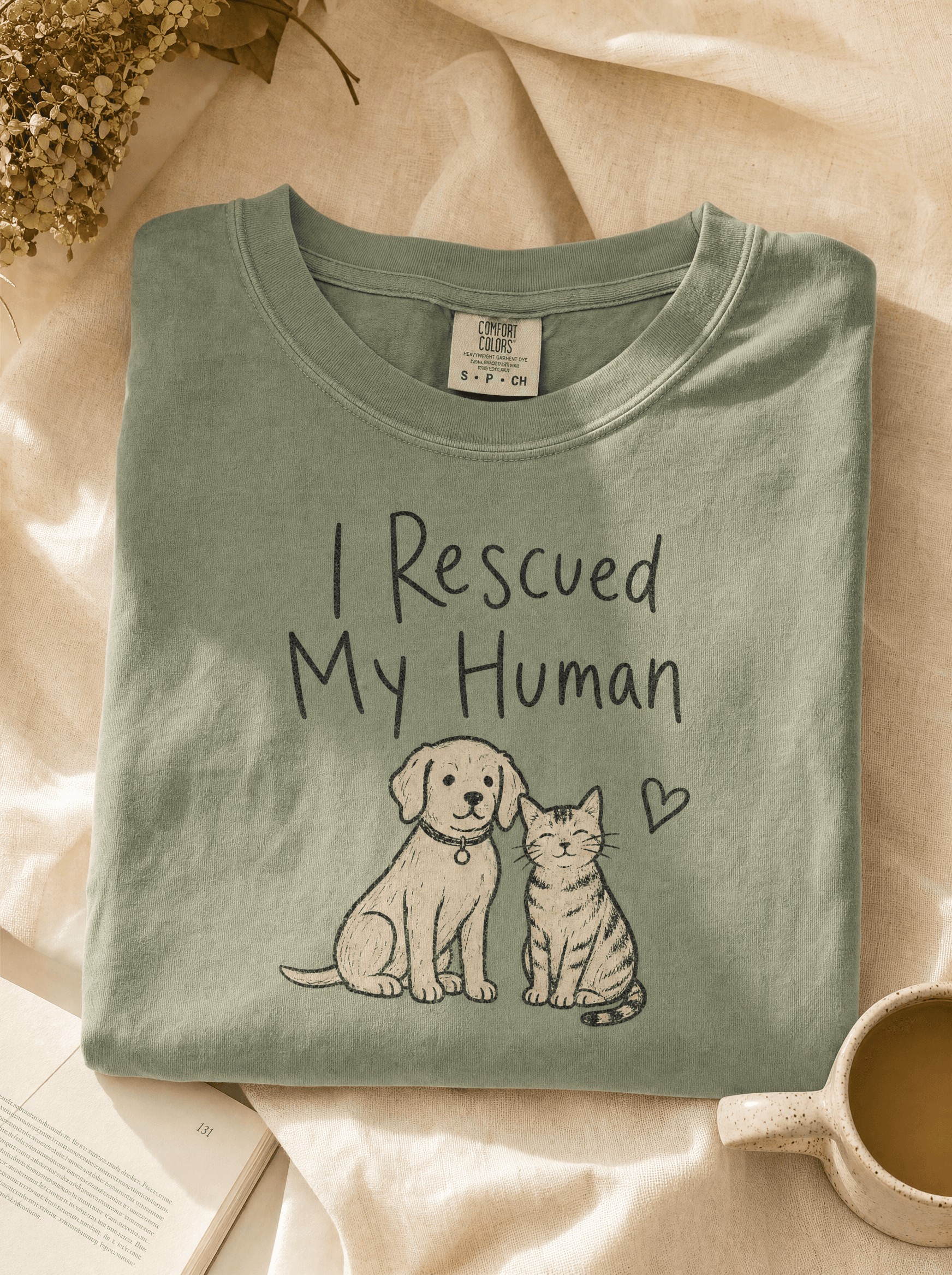 Heavyweight sage-green tee folded on cream linen with 'I Rescued My Human' typography